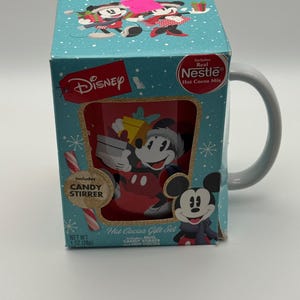May include: A Disney Hot Cocoa Gift Set featuring Mickey Mouse. The box is teal with snowflake designs and includes a mug, candy stirrer, and Nestle hot cocoa mix. The mug has a grey handle and a Mickey Mouse graphic.