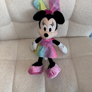 May include: A plush Minnie Mouse doll with black ears, a pink bow, and a colorful dress. The doll has white gloves and pink shoes. The dress features a rainbow gradient of colors.