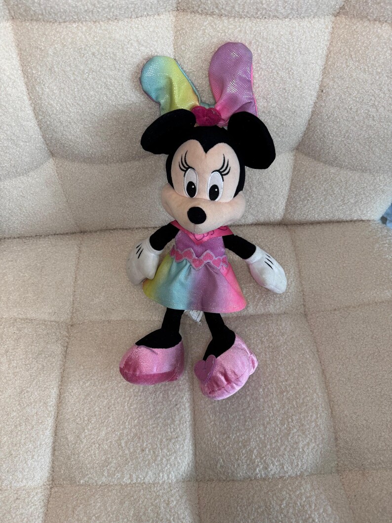 May include: A plush Minnie Mouse doll with black ears, a pink bow, and a colorful dress. The doll has white gloves and pink shoes. The dress features a rainbow gradient of colors.