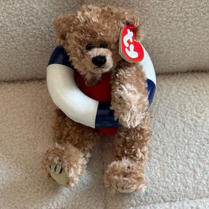 May include: A brown teddy bear wearing a red and blue life preserver. The plush toy has a black nose and a red tag attached to its ear. The bear is sitting on a white, textured surface.