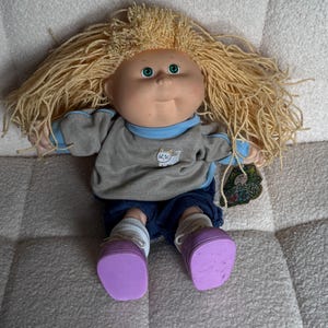 May include: A Cabbage Patch Kids doll with blonde yarn hair, green eyes, and a round face. It wears a gray sweatshirt with a cat graphic, blue jeans, purple shoes, and white socks. A tag is attached.
