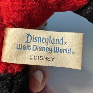 May include: A close-up of a vintage plush toy with a red body and black accents. A rectangular tag features the text "Disneyland Walt Disney World &copy;DISNEY" in blue lettering.