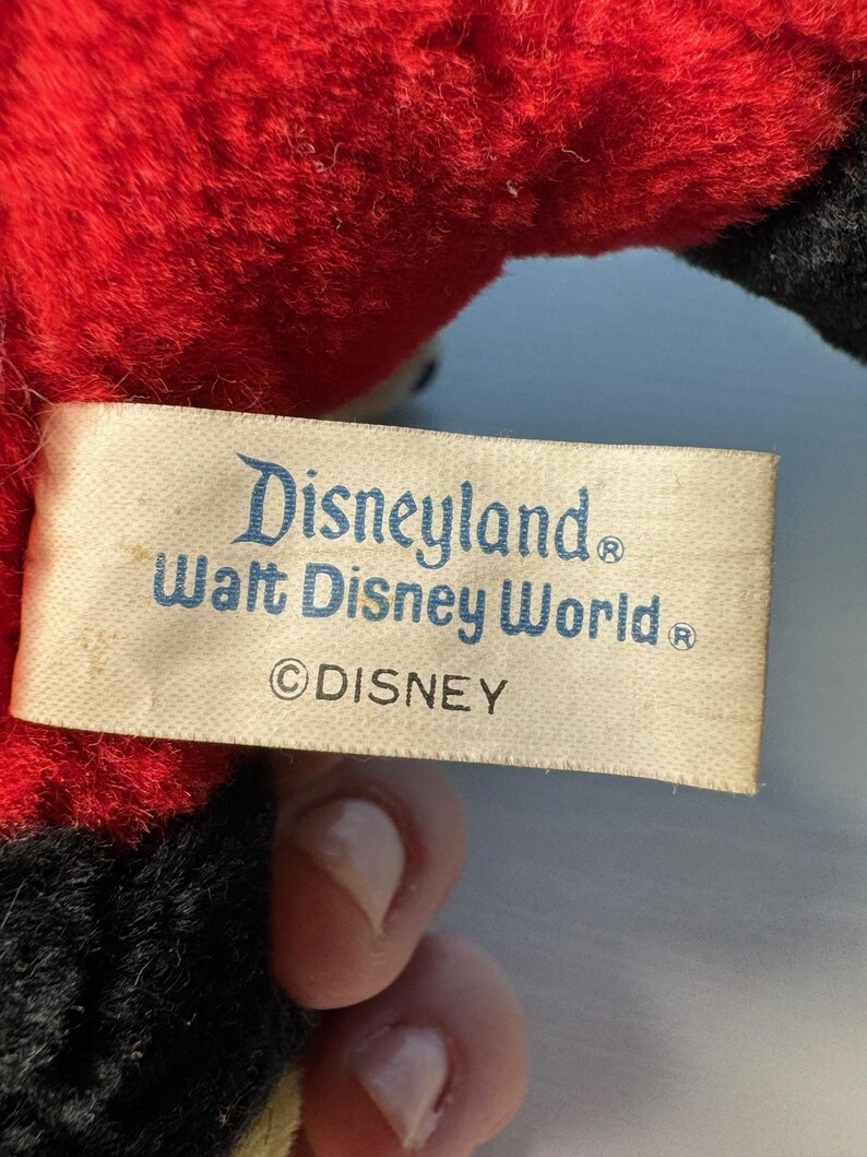 May include: A close-up of a vintage plush toy with a red body and black accents. A rectangular tag features the text "Disneyland Walt Disney World &copy;DISNEY" in blue lettering.