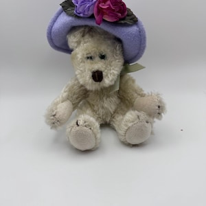 May include: A plush teddy bear wearing a light purple hat adorned with purple and magenta fabric flowers. The bear has a light brown nose and green eyes. A green ribbon is tied around the bear's neck.