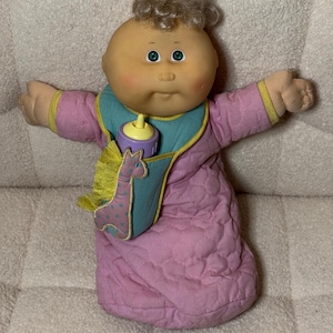 May include: A vintage Cabbage Patch Kids doll with a soft, round face, blue eyes, and light brown curly hair. The doll wears a pink quilted outfit with yellow trim and a teal bib with a giraffe design and a bottle.