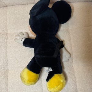 Puede incluir: Un peluche de Mickey Mouse negro con pies amarillos y guantes blancos. El mu&ntilde;eco tiene orejas grandes y redondas y una expresi&oacute;n sonriente. Este personaje cl&aacute;sico es un objeto de colecci&oacute;n popular.