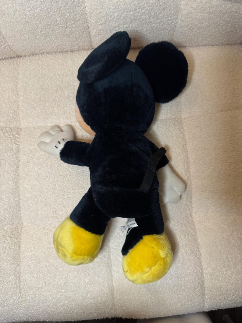 Puede incluir: Un peluche de Mickey Mouse negro con pies amarillos y guantes blancos. El mu&ntilde;eco tiene orejas grandes y redondas y una expresi&oacute;n sonriente. Este personaje cl&aacute;sico es un objeto de colecci&oacute;n popular.