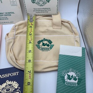 May include: A cream-coloured canvas bag with Cabbage Patch Kids Airways logo, accompanied by a passport, a card, and a green envelope. A yellow Stanley measuring tape shows the bag is approximately 19 cm tall. The items feature green text.