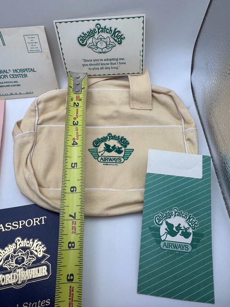 May include: A cream-coloured canvas bag with Cabbage Patch Kids Airways logo, accompanied by a passport, a card, and a green envelope. A yellow Stanley measuring tape shows the bag is approximately 19 cm tall. The items feature green text.