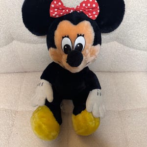 Puede incluir: Mu&ntilde;eca de peluche de Minnie Mouse con pelaje negro, orejas grandes y redondas y un lazo rojo con lunares blancos. La mu&ntilde;eca tiene una cara bronceada, guantes blancos y zapatos amarillos. Es un juguete infantil cl&aacute;sico.