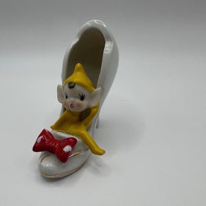 May include: A white ceramic shoe-shaped planter with a red bow and a yellow-clad elf figurine. The elf has a yellow hat and is resting inside the shoe. A decorative item.