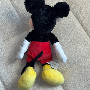 May include: A plush Mickey Mouse toy, viewed from the back, features black fur, red shorts, yellow shoes, and white gloves. The toy has a tag that reads "Disneyland, or Disney World".