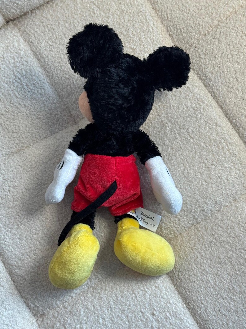 May include: A plush Mickey Mouse toy, viewed from the back, features black fur, red shorts, yellow shoes, and white gloves. The toy has a tag that reads "Disneyland, or Disney World".