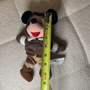 K&ouml;nnte beinhalten: Ein Pl&uuml;schtier von Mickey Mouse, etwa 20 cm gro&szlig;, in einem braunen Fliegeroutfit mit Kapuze. Das Spielzeug hat schwarze Ohren, einen roten Mund und wei&szlig;e Handschuhe. Ein gelbes Ma&szlig;band liegt neben dem Spielzeug.