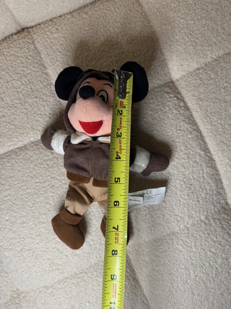 K&ouml;nnte beinhalten: Ein Pl&uuml;schtier von Mickey Mouse, etwa 20 cm gro&szlig;, in einem braunen Fliegeroutfit mit Kapuze. Das Spielzeug hat schwarze Ohren, einen roten Mund und wei&szlig;e Handschuhe. Ein gelbes Ma&szlig;band liegt neben dem Spielzeug.