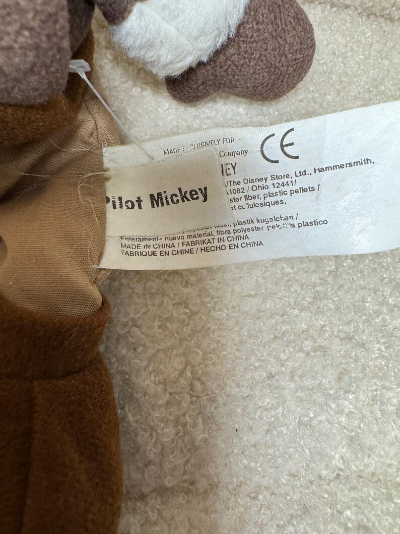 K&ouml;nnte beinhalten: Nahaufnahme eines Pilot Mickey Mouse Pl&uuml;schtiers. Das Spielzeug ist braun, wei&szlig; und beige. Auf dem Etikett steht "Pilot Mickey" und enth&auml;lt das CE-Zeichen. Das Etikett enth&auml;lt auch die Informationen von The Disney Store, Ltd.