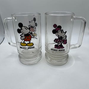 May include: Two clear glass mugs featuring Mickey Mouse and Minnie Mouse. Mickey is in his classic outfit, and Minnie is in a polka dot dress. Each mug has a handle and the character's name printed on the front. A vintage Disney collectible.