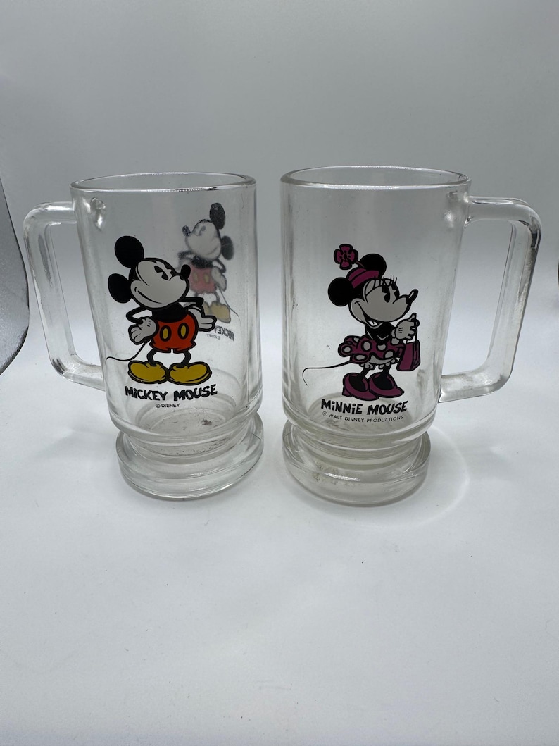 May include: Two clear glass mugs featuring Mickey Mouse and Minnie Mouse. Mickey is in his classic outfit, and Minnie is in a polka dot dress. Each mug has a handle and the character's name printed on the front. A vintage Disney collectible.