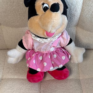 May include: A plush Minnie Mouse doll, featuring black ears, a pink bow, and a pink and white polka dot dress. The doll has white gloves and red shoes. The doll is sitting on a textured surface.