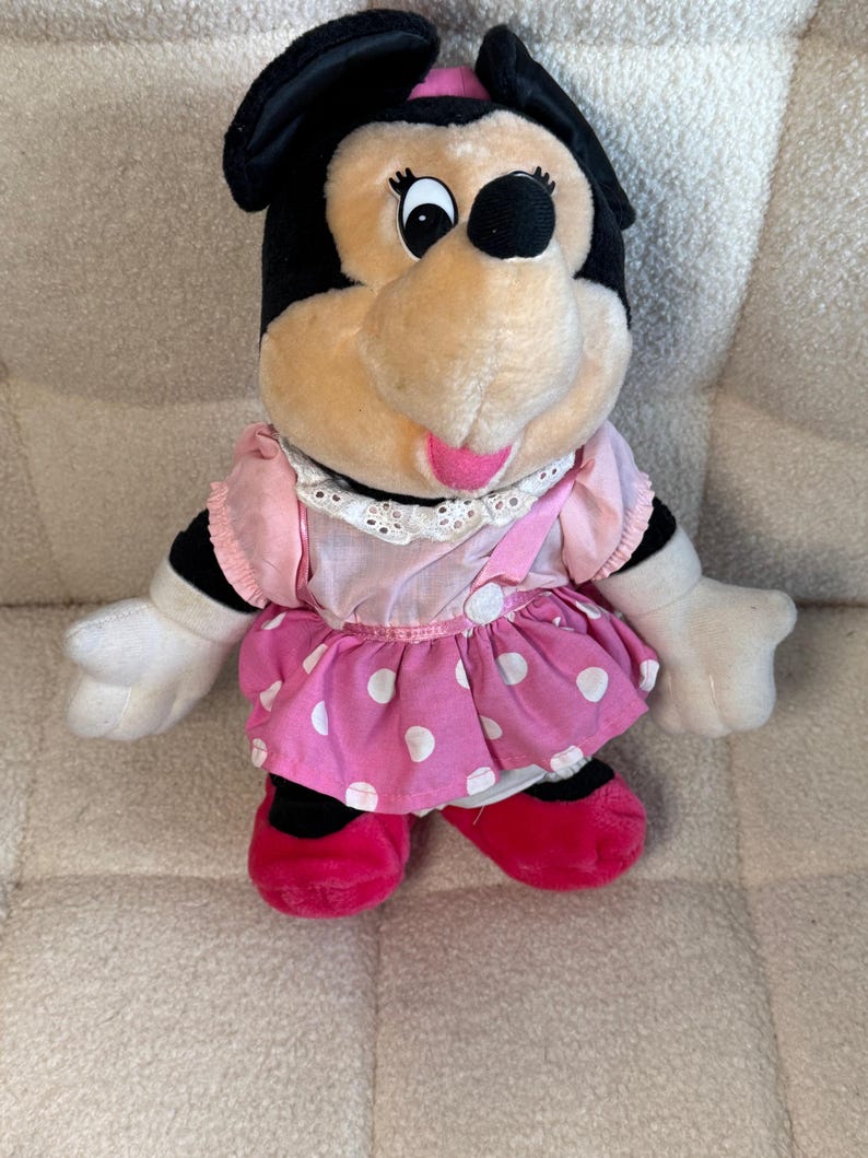 May include: A plush Minnie Mouse doll, featuring black ears, a pink bow, and a pink and white polka dot dress. The doll has white gloves and red shoes. The doll is sitting on a textured surface.