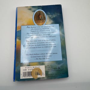 May include: The back cover of "The Black Stallion" book. The cover features a blue and tan background with text describing the story of a shipwreck survivor and a horse. A sticker with pricing information is on the bottom left.