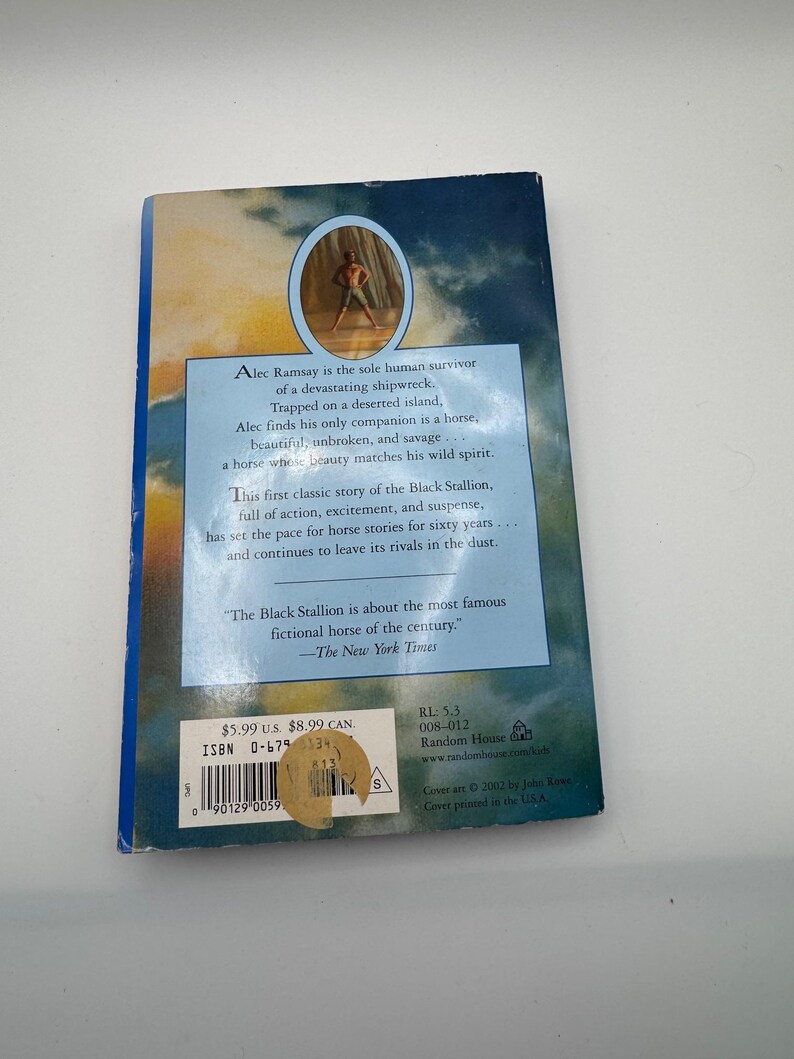 May include: The back cover of "The Black Stallion" book. The cover features a blue and tan background with text describing the story of a shipwreck survivor and a horse. A sticker with pricing information is on the bottom left.