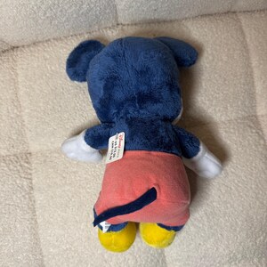May include: A plush Mickey Mouse toy, seen from the rear. The toy has a blue head and ears, white gloves, red shorts, yellow shoes, and a black tail. A Disney Store tag is attached.