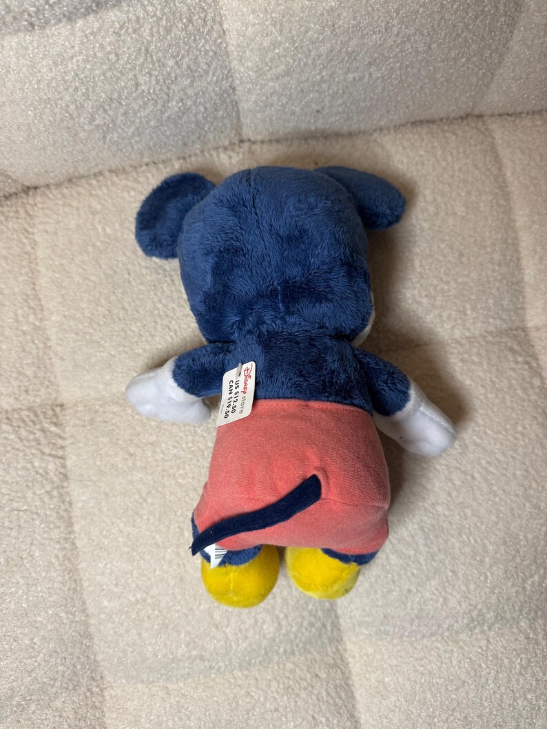 May include: A plush Mickey Mouse toy, seen from the rear. The toy has a blue head and ears, white gloves, red shorts, yellow shoes, and a black tail. A Disney Store tag is attached.