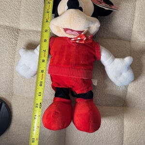 May include: A plush Mickey Mouse doll, approximately 16 inches tall, wearing a red sweater vest, shorts, and shoes. The doll has white gloves, a bow tie, and black ears. The doll is a classic Disney character.