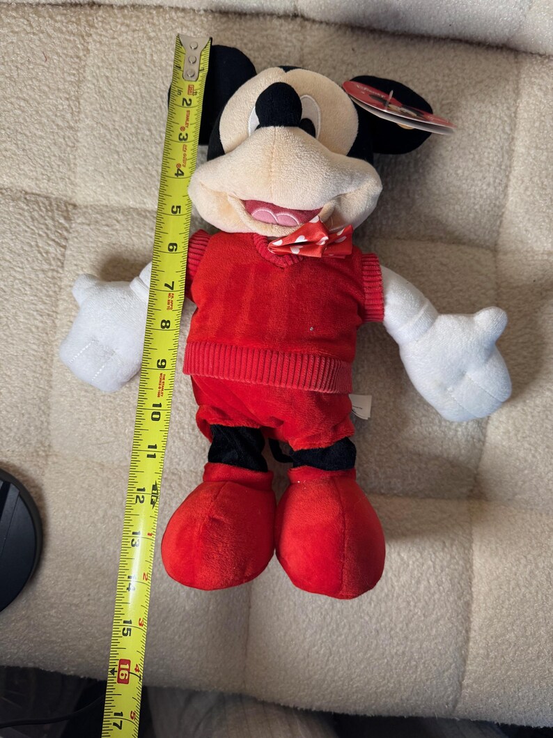 May include: A plush Mickey Mouse doll, approximately 16 inches tall, wearing a red sweater vest, shorts, and shoes. The doll has white gloves, a bow tie, and black ears. The doll is a classic Disney character.