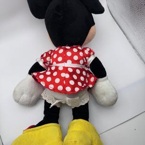 May include: A plush Minnie Mouse doll, viewed from the back, features a red dress with white polka dots, a matching bow, and large yellow shoes. The doll has black ears, arms, and legs, with white gloves. The doll is a classic children's toy.