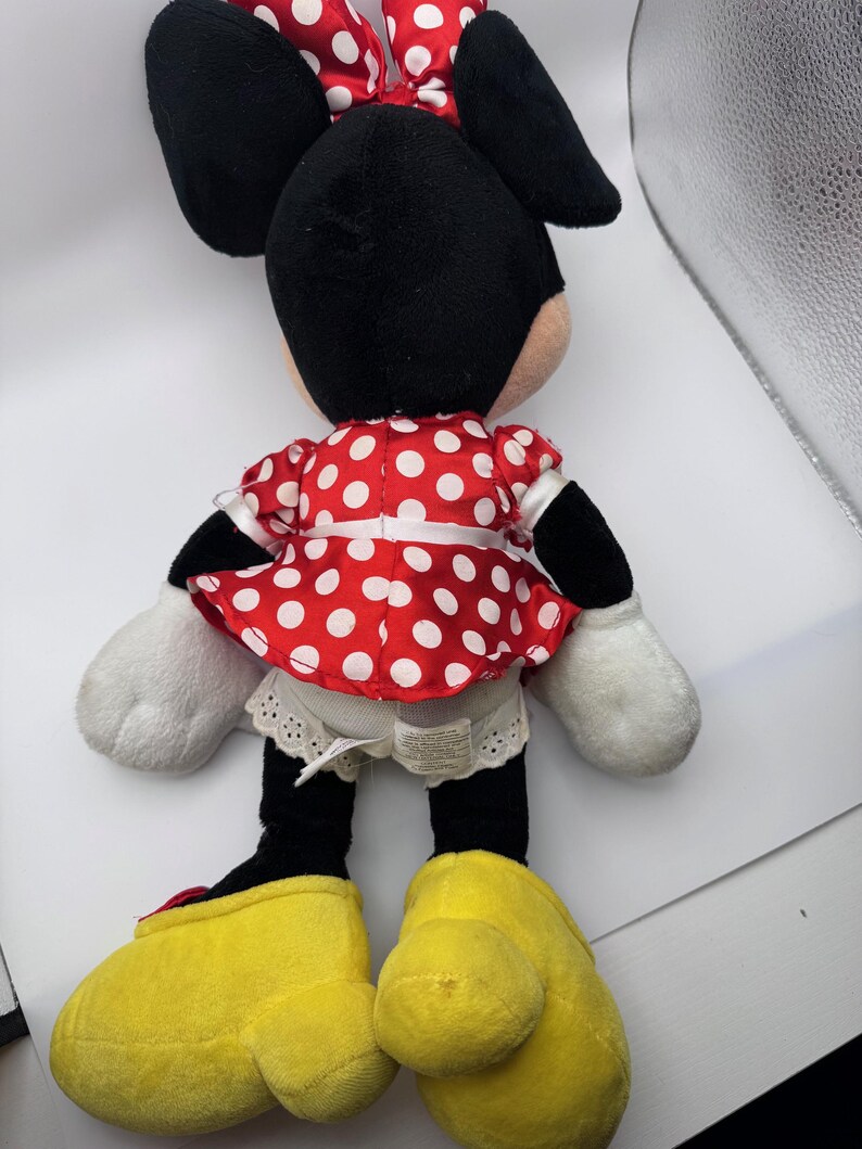 May include: A plush Minnie Mouse doll, viewed from the back, features a red dress with white polka dots, a matching bow, and large yellow shoes. The doll has black ears, arms, and legs, with white gloves. The doll is a classic children's toy.