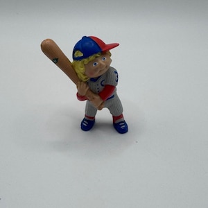 May include: A figurine of a baseball player in a batting stance. The player wears a blue and red cap, a gray uniform with the number 3, red sleeves, and blue shoes. The player is holding a wooden baseball bat.