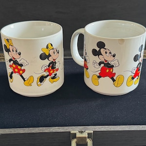May include: Two white ceramic mugs with handles. Both mugs feature a colorful cartoon design of Mickey Mouse and Minnie Mouse running. The mugs are on a dark surface.