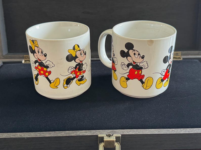 May include: Two white ceramic mugs with handles. Both mugs feature a colorful cartoon design of Mickey Mouse and Minnie Mouse running. The mugs are on a dark surface.