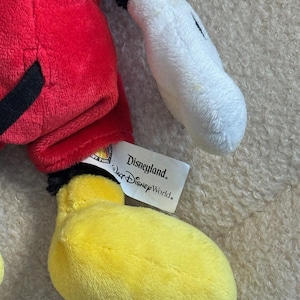 May include: A plush Mickey Mouse toy featuring a red shorts, black tail, yellow feet, and white gloves. The toy has a black head and a tag that reads "Disneyland Walt Disney World."