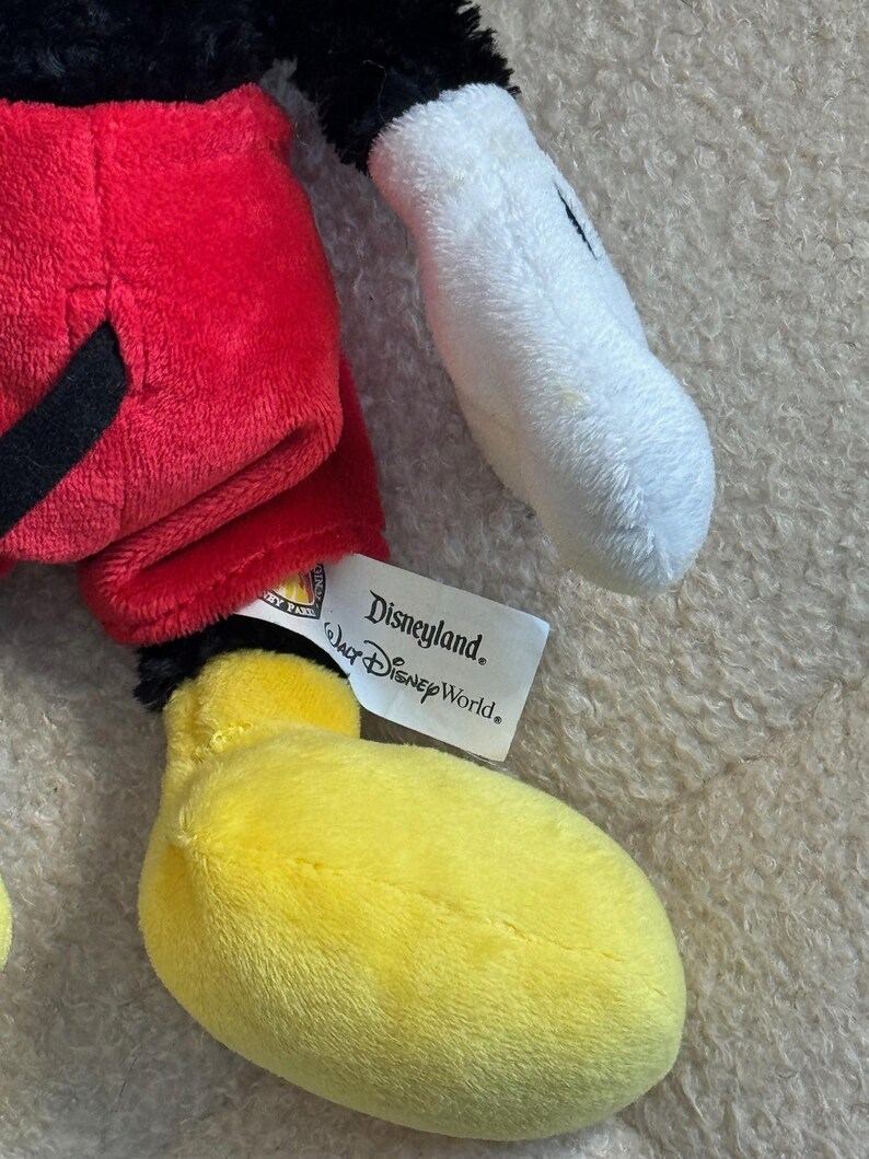 May include: A plush Mickey Mouse toy featuring a red shorts, black tail, yellow feet, and white gloves. The toy has a black head and a tag that reads "Disneyland Walt Disney World."