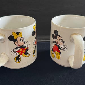May include: Two white ceramic mugs with handles. One mug features a cartoon image of Mickey Mouse and Minnie Mouse. The other mug features a cartoon image of Minnie Mouse and a smaller Minnie Mouse.