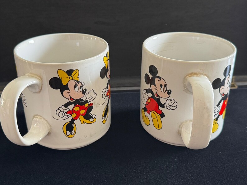 May include: Two white ceramic mugs with handles. One mug features a cartoon image of Mickey Mouse and Minnie Mouse. The other mug features a cartoon image of Minnie Mouse and a smaller Minnie Mouse.