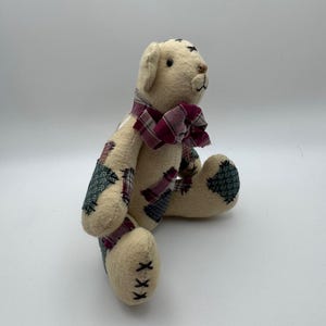 May include: A handmade teddy bear with a cream-colored body and patchwork accents in green, red, and blue. The bear has a burgundy plaid bow tie and black stitched details. The bear is sitting with its legs extended.