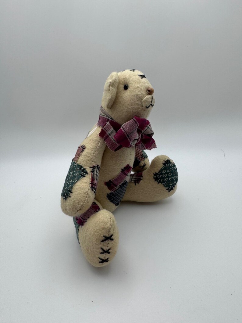 May include: A handmade teddy bear with a cream-colored body and patchwork accents in green, red, and blue. The bear has a burgundy plaid bow tie and black stitched details. The bear is sitting with its legs extended.
