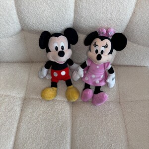 May include: Two plush Disney characters, Mickey Mouse and Minnie Mouse, are displayed. Mickey wears red shorts, yellow shoes, and black ears. Minnie is in a pink dress with white polka dots, pink shoes, and a pink bow. Both have white gloves.
