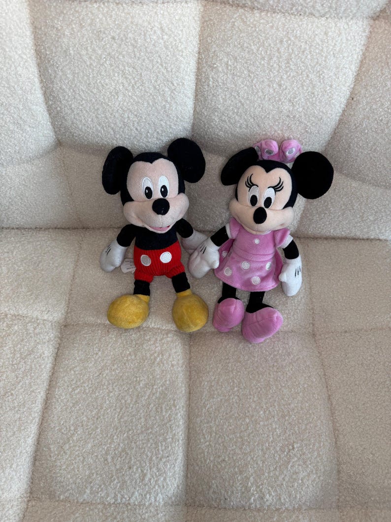 May include: Two plush Disney characters, Mickey Mouse and Minnie Mouse, are displayed. Mickey wears red shorts, yellow shoes, and black ears. Minnie is in a pink dress with white polka dots, pink shoes, and a pink bow. Both have white gloves.