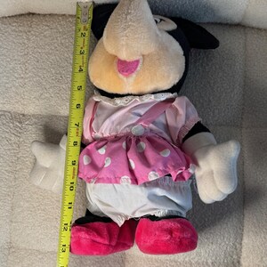 May include: A plush Minnie Mouse doll, approximately 14 inches tall, with a black head and ears, a pink and white polka dot skirt, and pink shoes. The doll has white gloves and a pink bow.