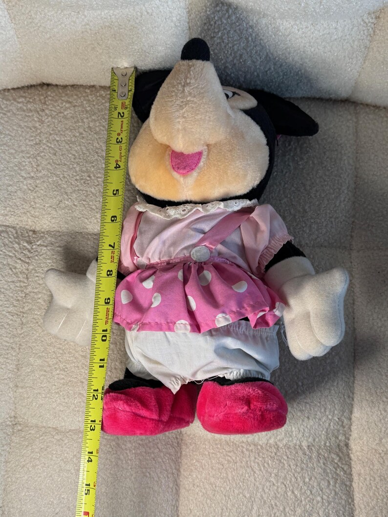 May include: A plush Minnie Mouse doll, approximately 14 inches tall, with a black head and ears, a pink and white polka dot skirt, and pink shoes. The doll has white gloves and a pink bow.