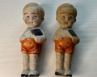 May include: Two antique bisque dolls of young children. Each doll is dressed in orange shorts, white shirts, and black shoes. They are holding a black book. The dolls have blonde hair and are standing upright.