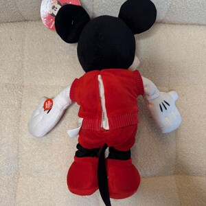 May include: A plush Mickey Mouse doll, viewed from the back, with black ears, a black tail, and red shorts and jacket. The jacket has a zipper down the back. White gloves and a tag are also visible.