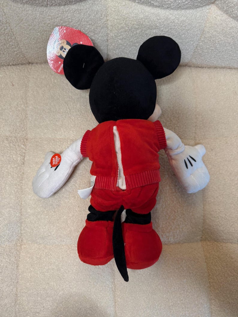 May include: A plush Mickey Mouse doll, viewed from the back, with black ears, a black tail, and red shorts and jacket. The jacket has a zipper down the back. White gloves and a tag are also visible.