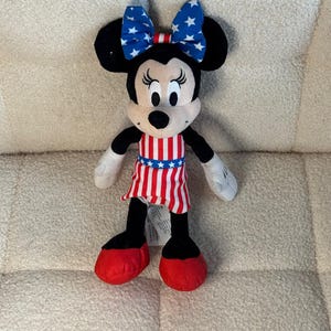 May include: A Minnie Mouse soft toy with black ears, a white face, and a red and white striped dress. The bow on her head is blue with white stars. Her shoes are red, and her arms and legs are black and white.