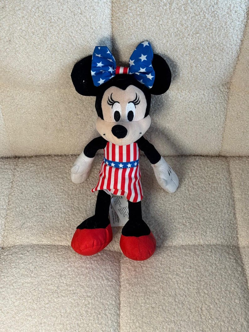 May include: A Minnie Mouse soft toy with black ears, a white face, and a red and white striped dress. The bow on her head is blue with white stars. Her shoes are red, and her arms and legs are black and white.